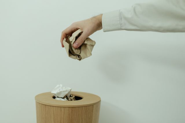 A person's hand disposes of crumpled paper into a wooden trash bin indoors.
